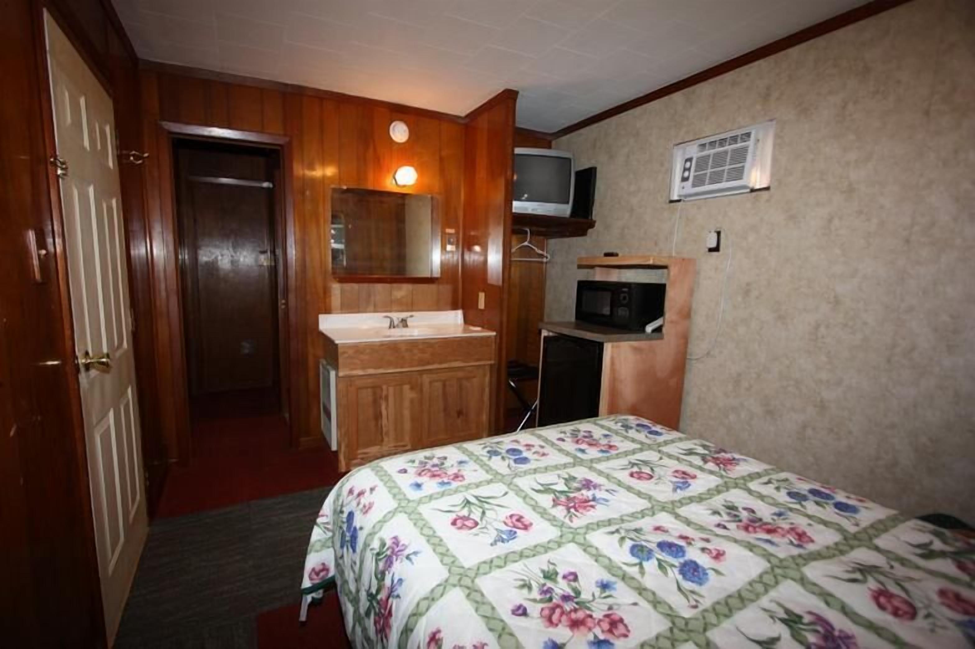 A hotel room with a wooden door, a sink, a mirror, a television, a microwave, and a bed with a floral bedspread.