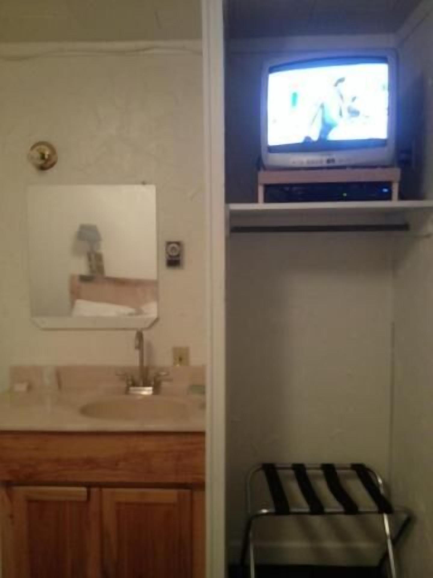 A bathroom with a wooden vanity, a mirror, a sink, a television on a shelf, and a towel rack.