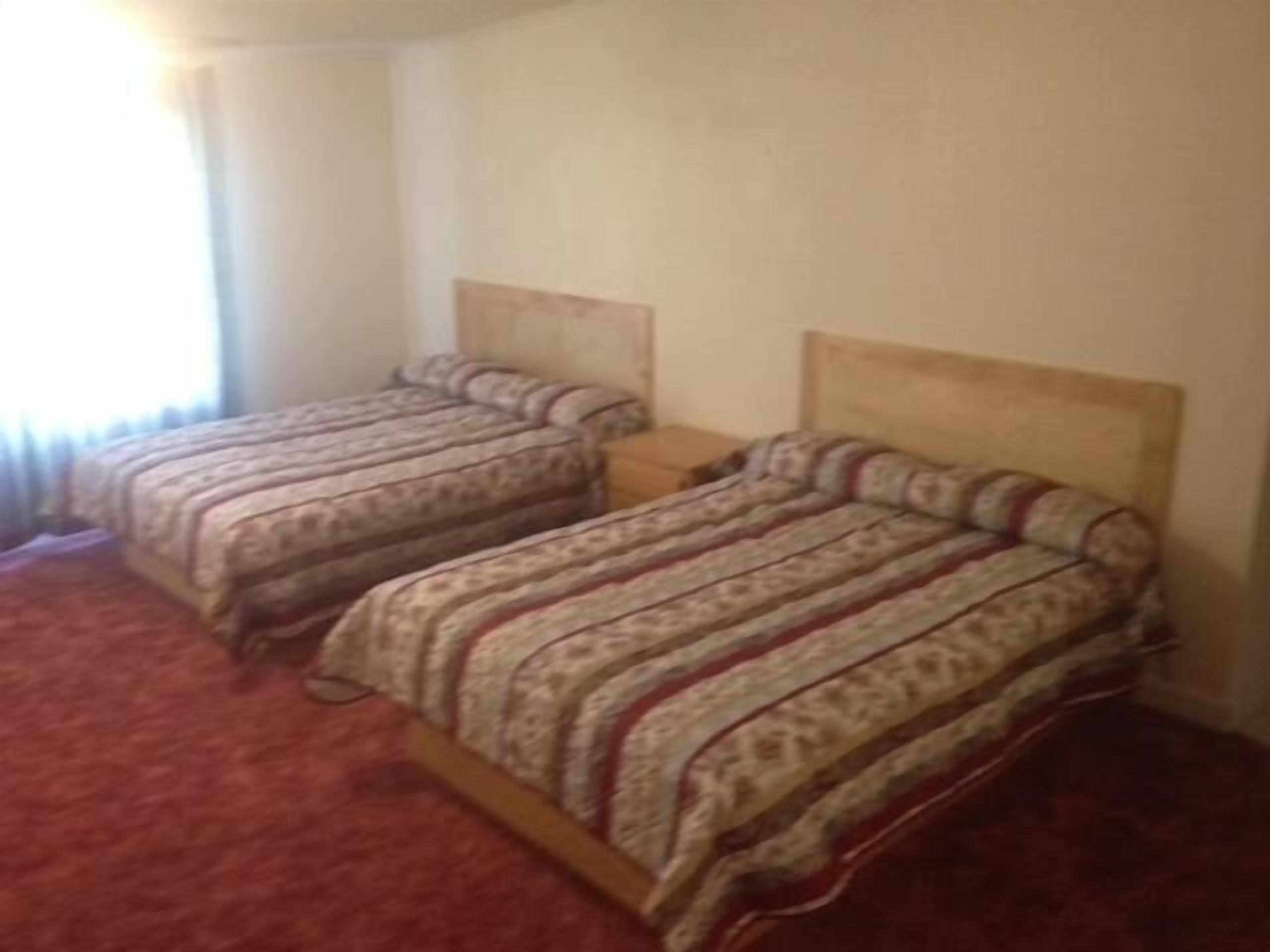 A room with two single beds, each with a wooden headboard, a small bedside table, and a red carpet.