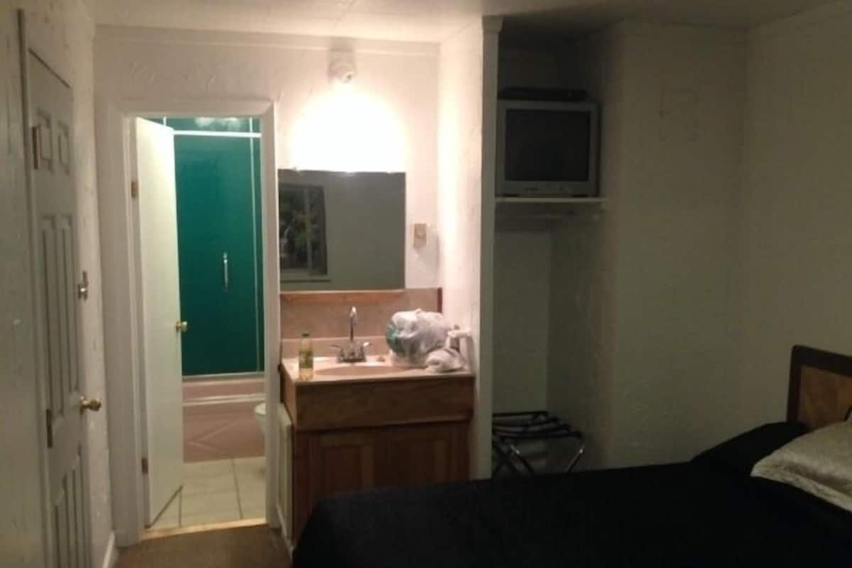 A small, dimly lit room with a single bed, a wooden dresser, a mirror, and a green door.