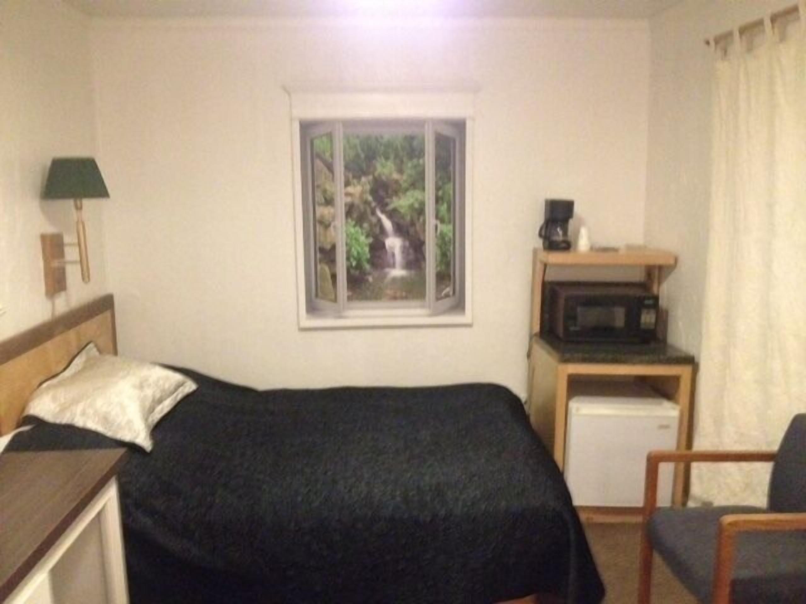 A bedroom with a bed, a desk with a microwave, a chair, a window with a view of a waterfall, and a lamp on the left.