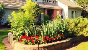 A two-story house with a tiled roof, a brick chimney, and a front porch with a small garden featuring red flowers and green plants. - McDougall Lane Bed & Breakfast (Drumheller)