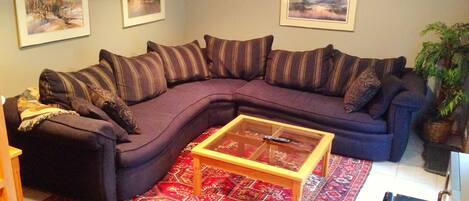 Ein Wohnzimmer mit einem dunkelgrauen Ecksofa, einem Glastisch, gemusterten Teppichen und drei gerahmten Landschaftsbildern an der Wand.