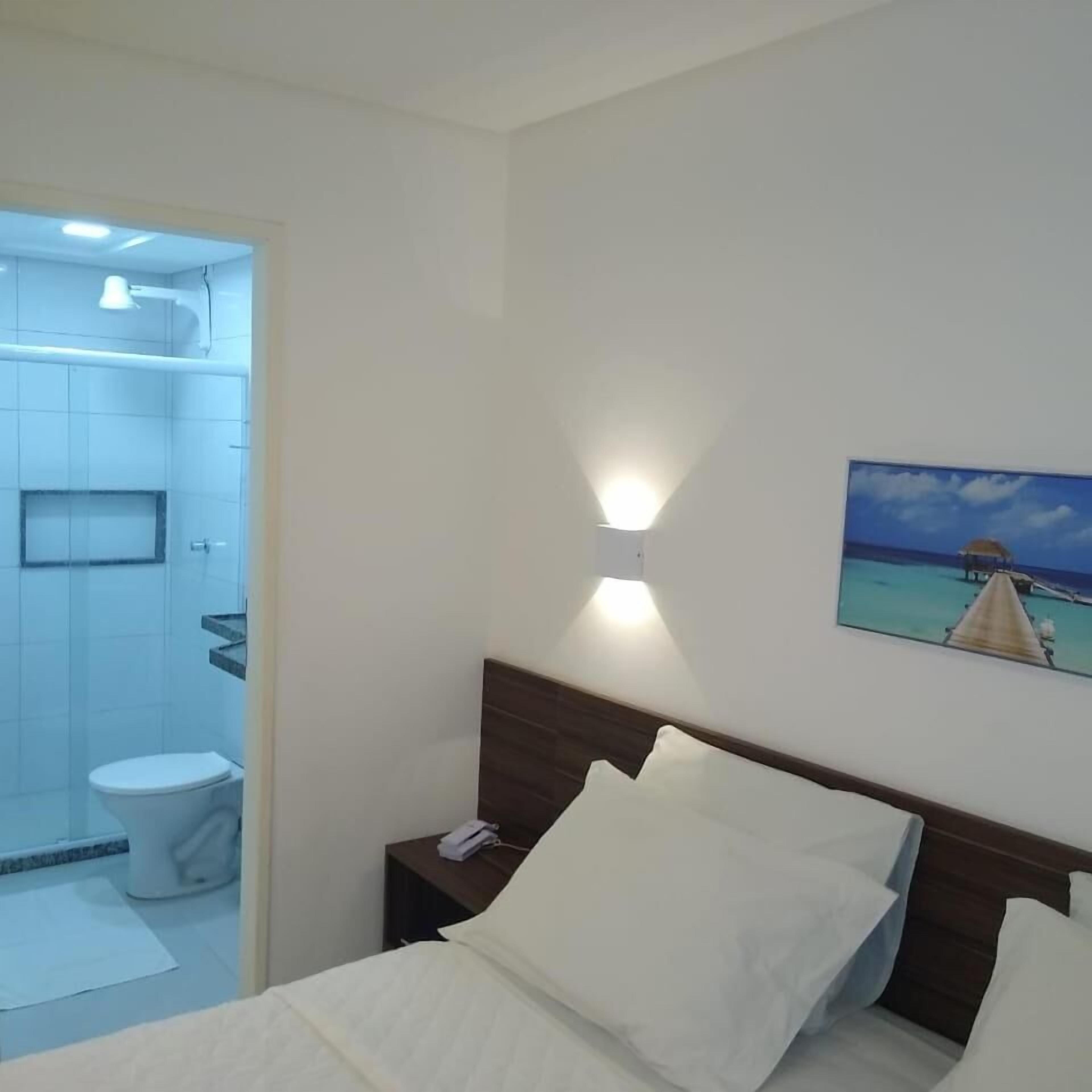A hotel room with a bed, a nightstand, a painting of a beach scene, and a bathroom with a toilet and shower.