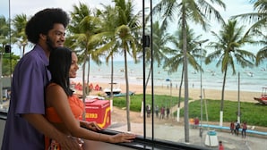 A couple standing by a window overlooking a beach with palm trees and boats. - Pousada Galemares (Maragogi)