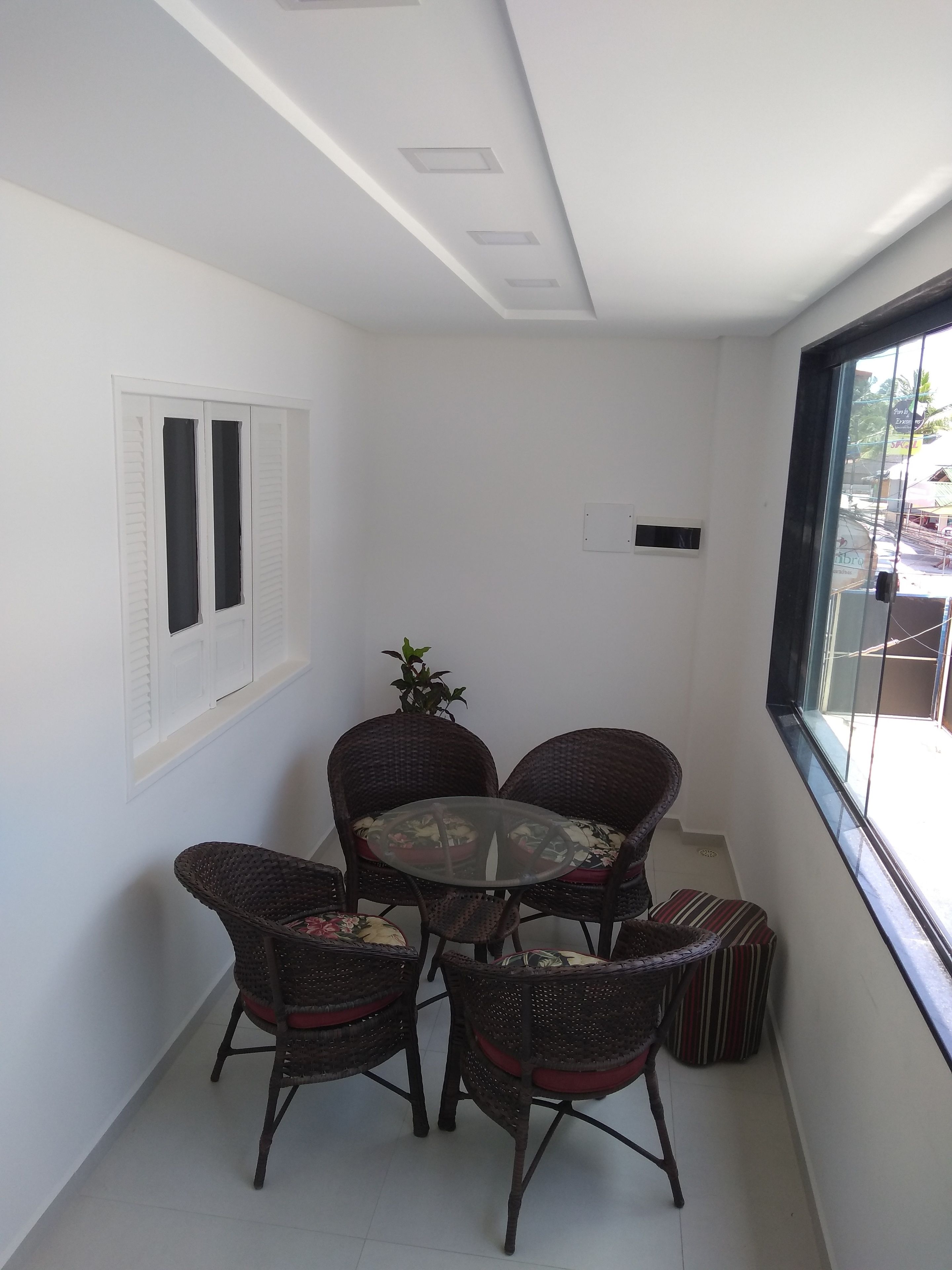 A small, enclosed balcony with a round table and four wicker chairs, a potted plant, and a window with blinds.