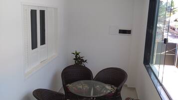 A small, enclosed balcony with a round table and four wicker chairs, a potted plant, and a window with blinds.