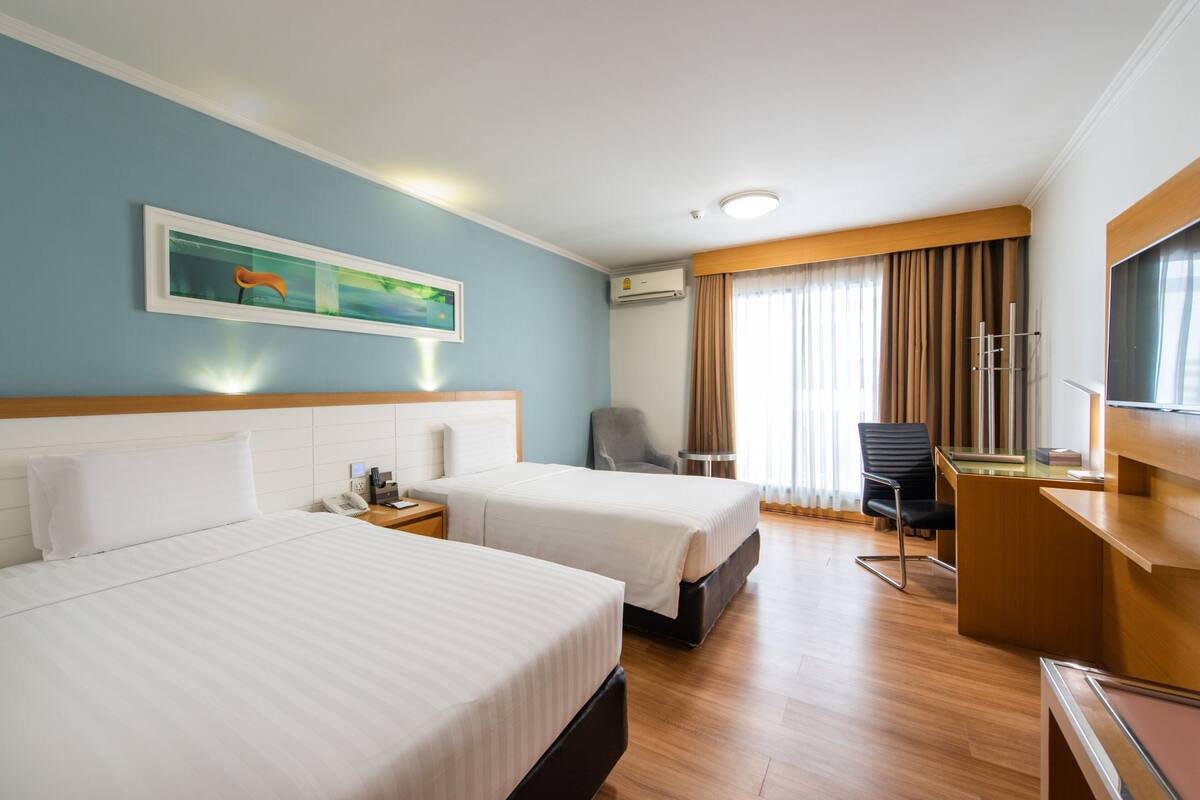 A hotel room with two beds, a desk, a chair, an air conditioning unit, and a painting on the wall.