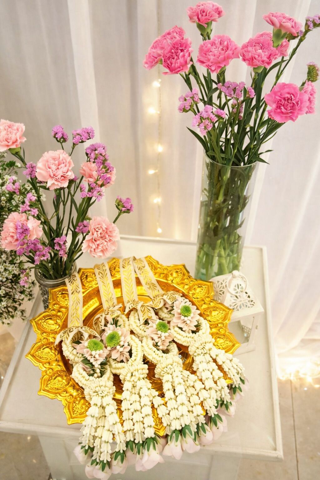 A table with a golden decorative tray holding white and pink floral garlands, pink carnations in a vase, and a small white decorative item.