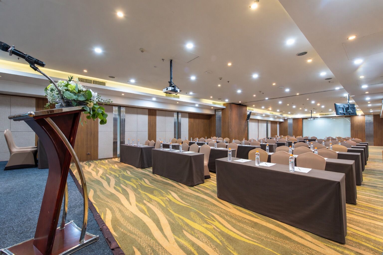 Une salle de conférence avec des rangées de tables et de chaises, un pupitre équipé d’un microphone et un écran de projection.