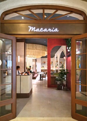 Una zona de recepció amb el rètol 'Macaria', una taquilla d'atenció al client, una zona de seients i un passadís amb plantes en testos.