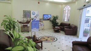 A lobby with a tiled floor, a coffee table, two brown sofas, a potted plant, a television, and a poster on the wall. - Alferdous Hotel Apartments (Muscat)