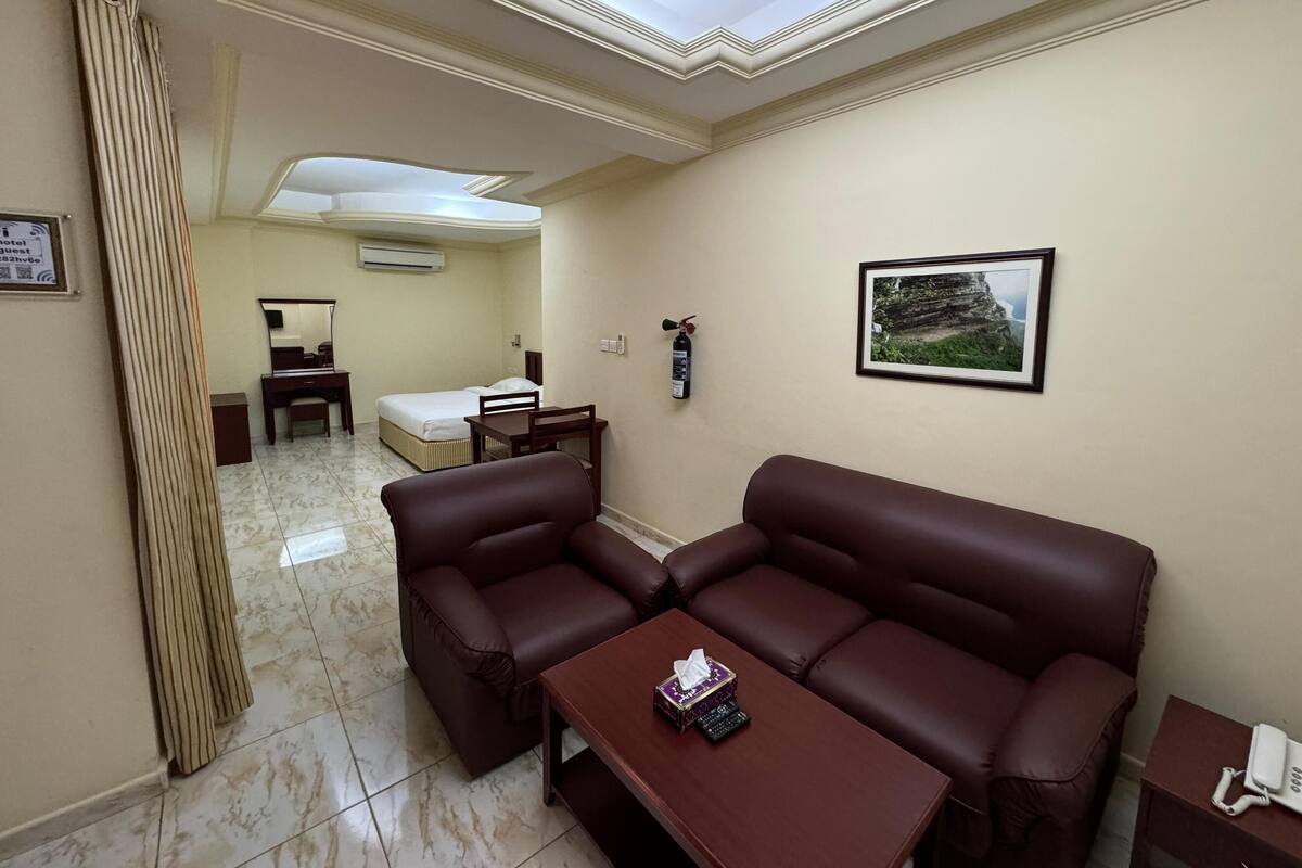 A hotel room with a bed, a desk, a chair, a sofa, a table, a picture on the wall, and a fire extinguisher.