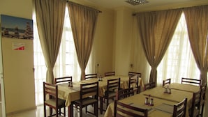 A dining area with multiple tables and chairs, large windows with curtains, and a tiled floor. - Alferdous Hotel Apartments (Muscat)