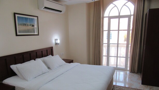 A neatly made bed in a hotel room with a large window, a wall-mounted air conditioner, and a framed picture on the wall. - Alferdous Hotel Apartments (Muscat)