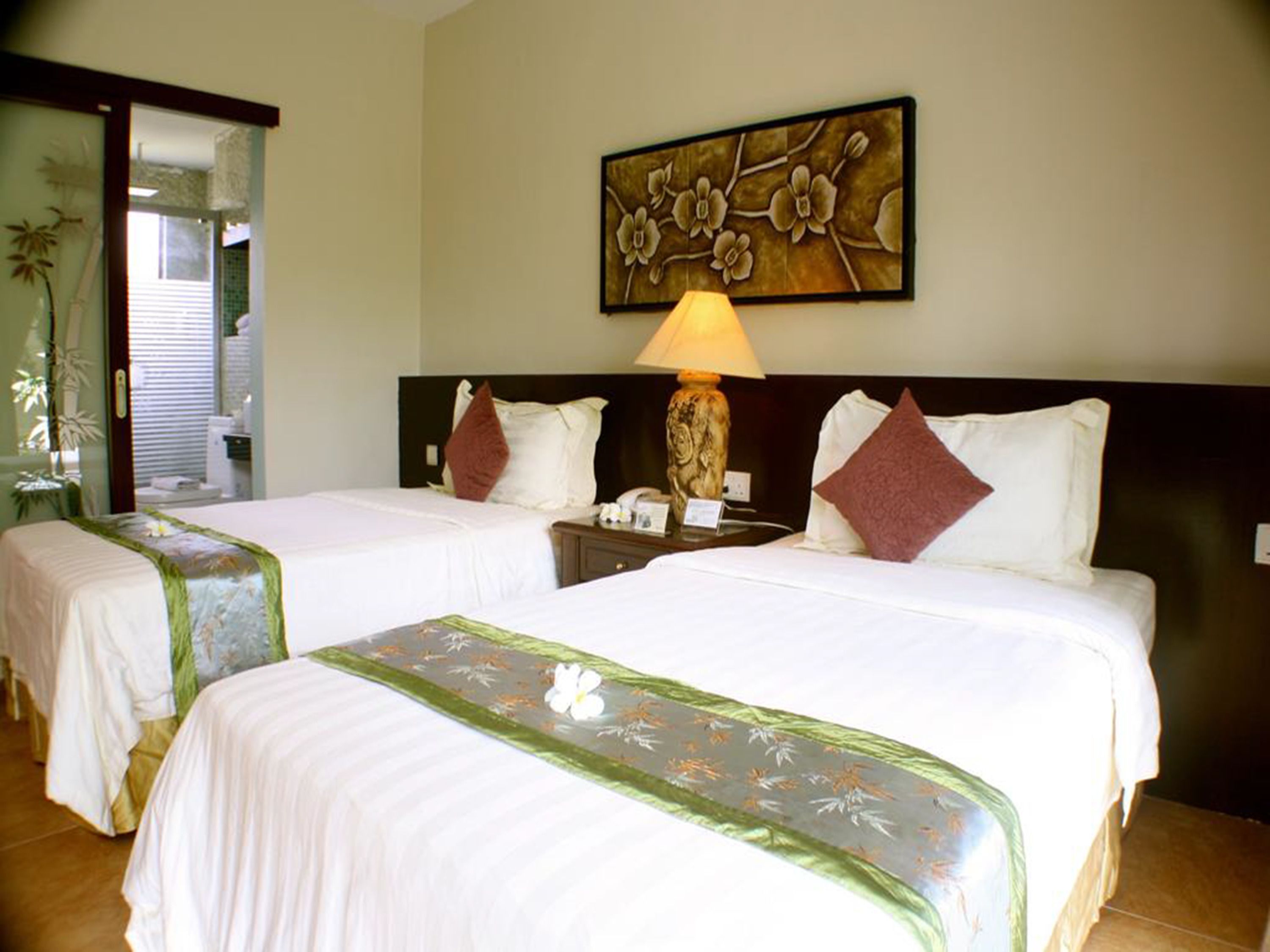 A hotel room with two beds, a nightstand, a lamp, and a floral artwork on the wall.