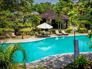 A swimming pool with lounge chairs and umbrellas, surrounded by lush greenery and a building in the background. - Beringgis Beach Resort & Spa (Papar)