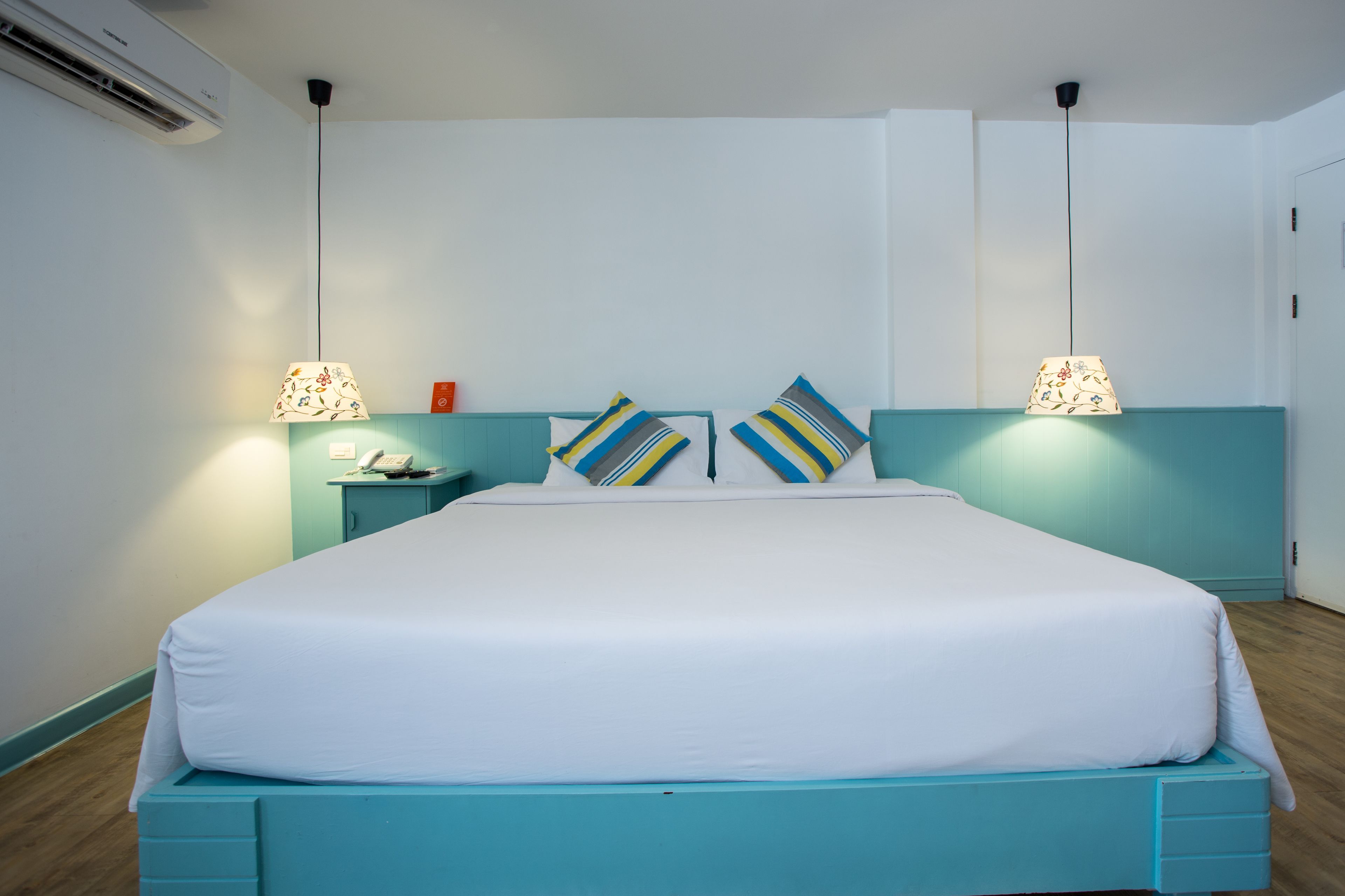 A neatly made bed with white bedding and blue headboard, two bedside lamps with floral lampshades, and a small bedside table with a phone and a red object.