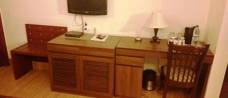 A compact room with a wooden desk, a chair, a television mounted on the wall, a lamp, and a trash can.
