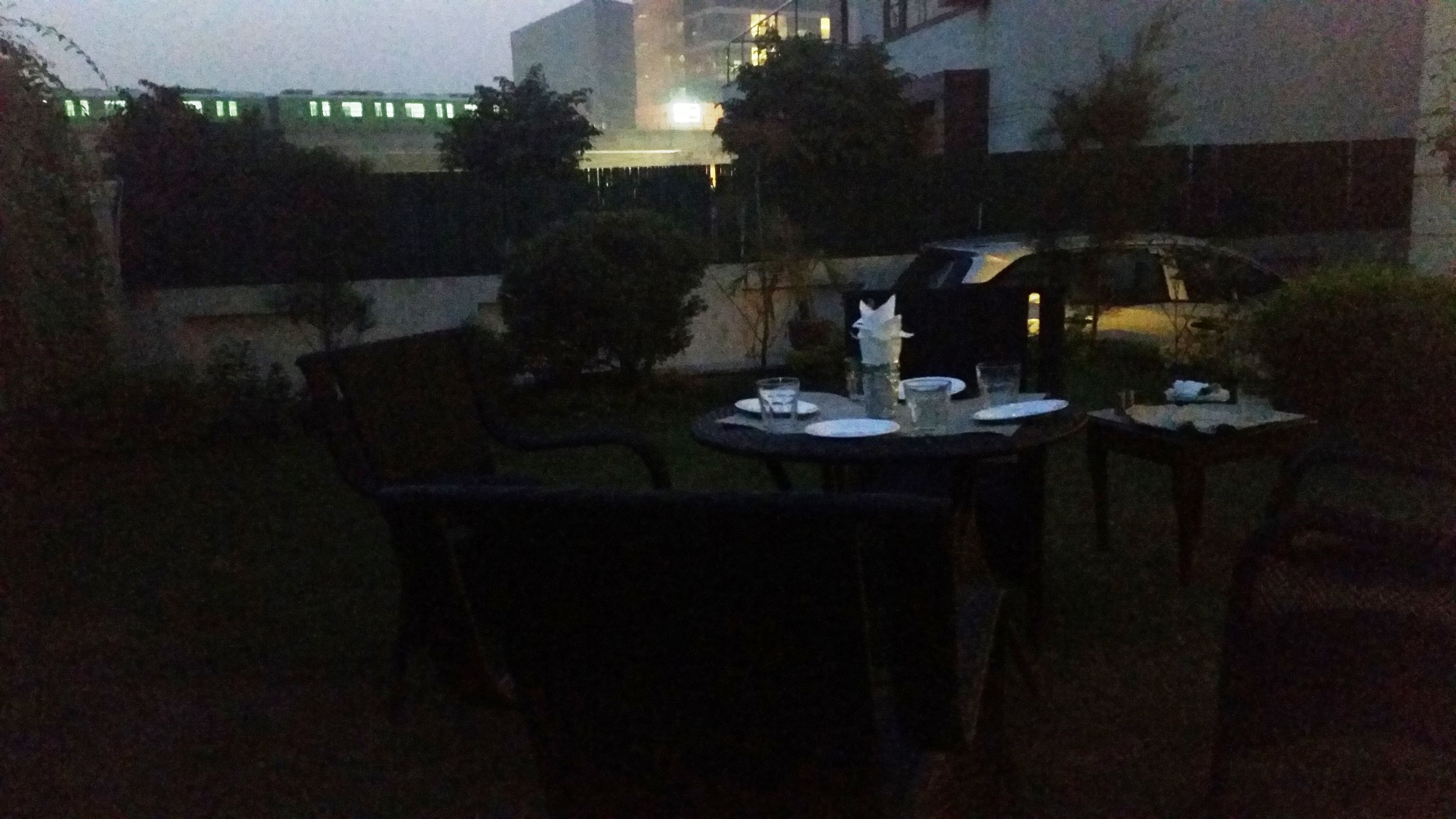 An outdoor dining area with tables and chairs, a small table with a bottle and glasses, and a building in the background.