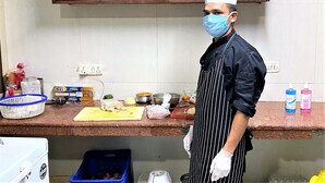 A kitchen with wooden cabinets, a countertop with various food items and kitchenware, and a person wearing a chef's hat and apron. - Tavisha Villa (Gurugram)