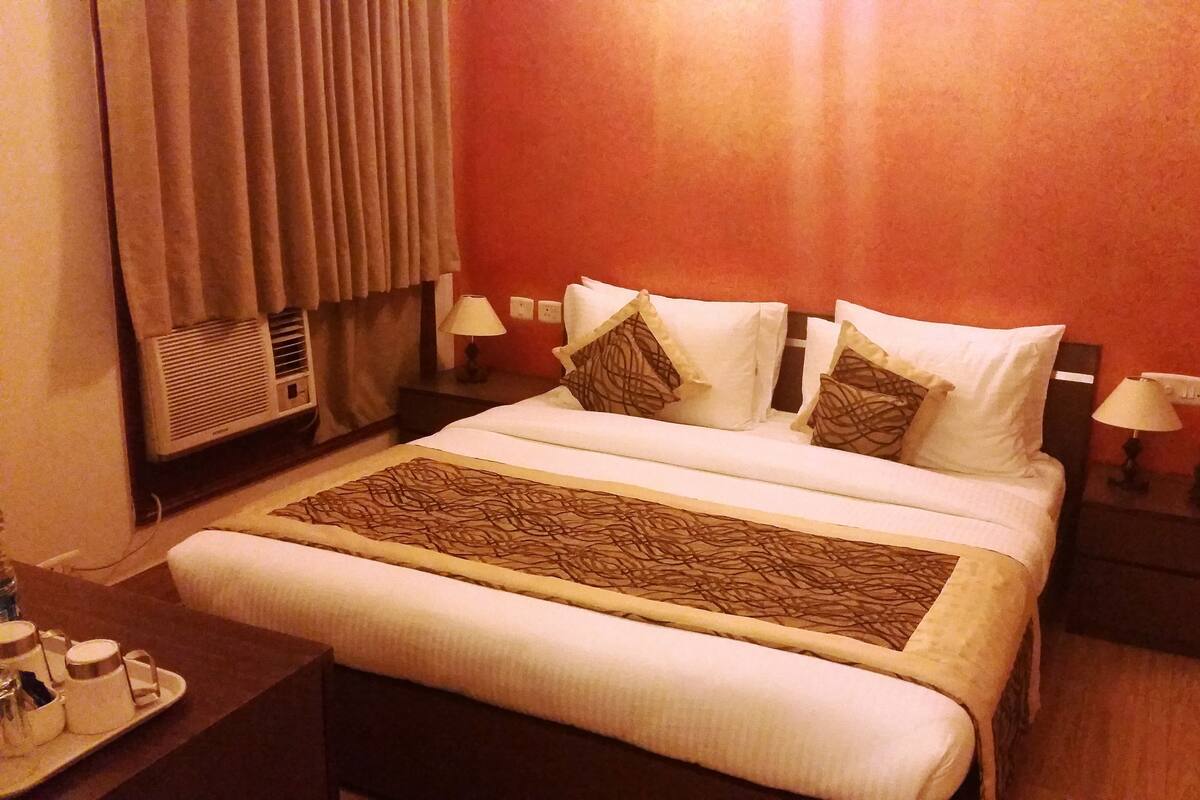 Una habitación de hotel con una cama grande, una mesita de noche con lámpara, un calefactor y una bandeja con tazas y una botella.