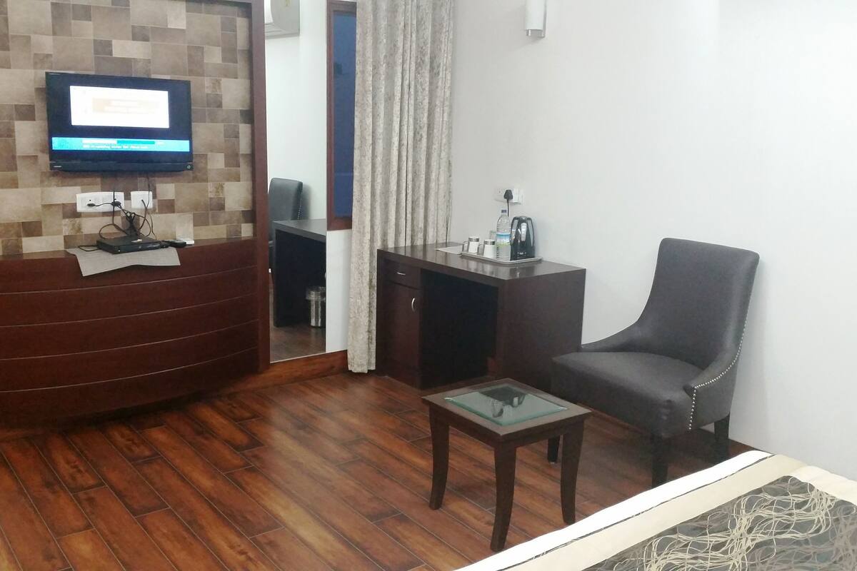 Una sala de estar con piso de madera, un televisor de pantalla plana montado en una pared de azulejos, un sofá marrón oscuro, una pequeña mesa de vidrio y un mueble de madera color café.