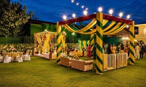Een evenementenlocatie buiten, met tafels en stoelen, een podium met bloemdecoraties, en een menigte mensen.