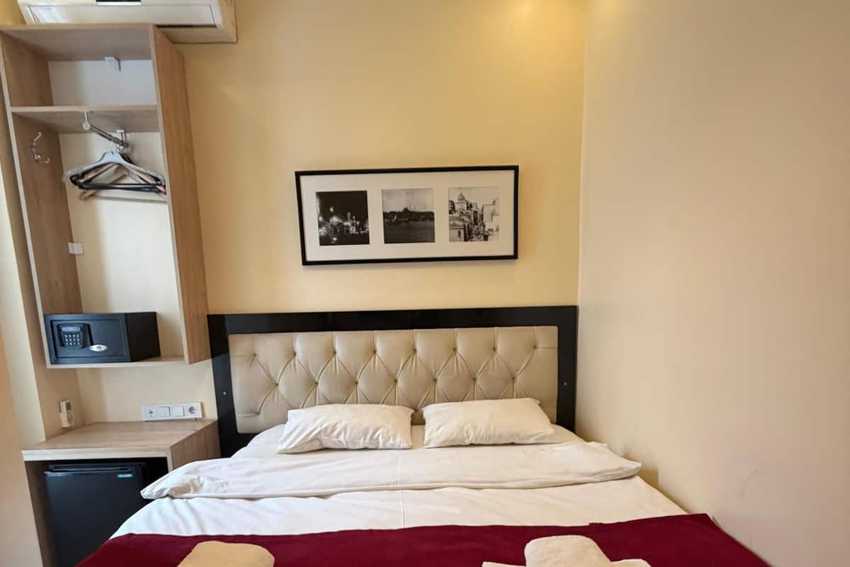 Ein ordentlich bezogenes Bett mit roter Decke, ein Nachttisch mit Lampe, eine Wandklimaanlage und ein gerahmtes Foto über dem Bett.