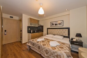 A bedroom with a bed, a nightstand, a lamp, a kitchenette, and a door.