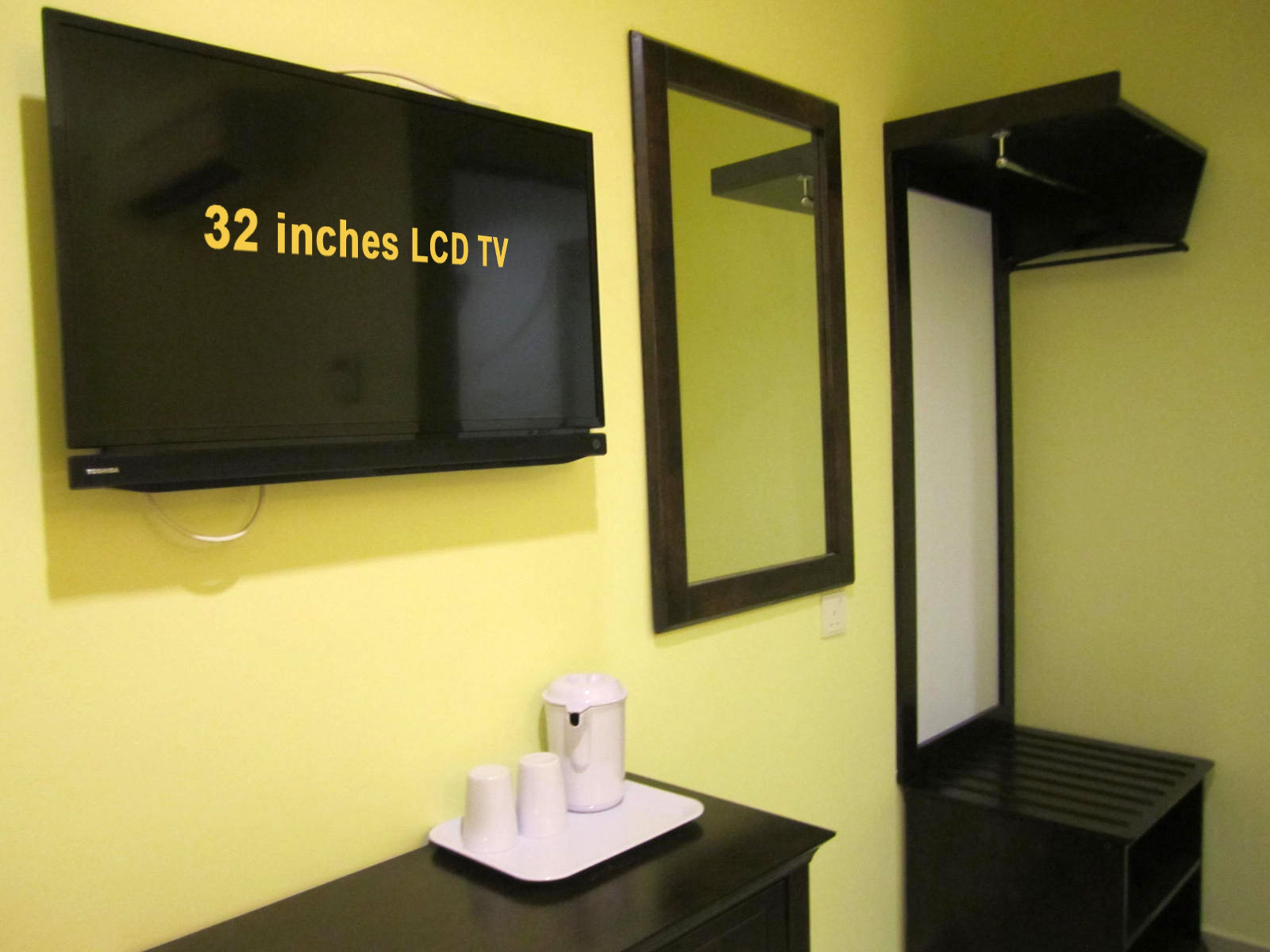 Un televisor LCD de 32 pulgadas montado en una pared amarilla, un espejo y una mesita con una cafetera y tazas.