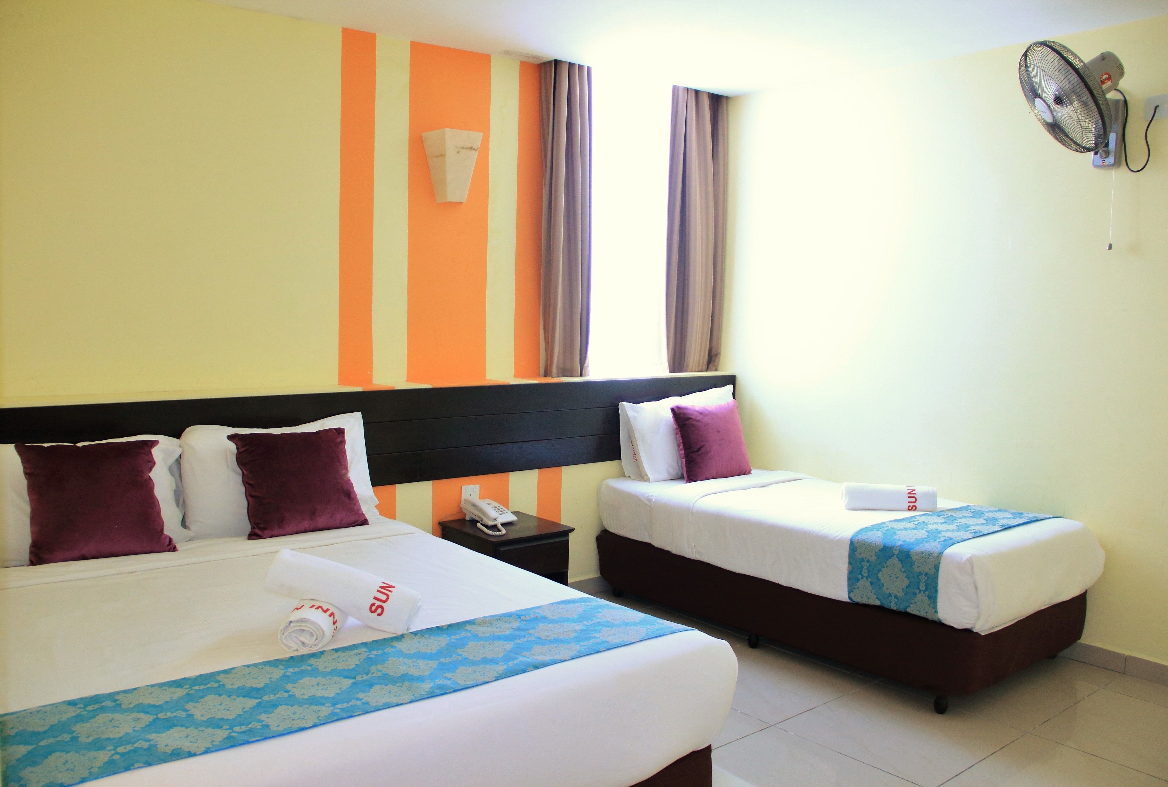 A hotel room with two beds, a nightstand with a phone, and a wall-mounted fan.