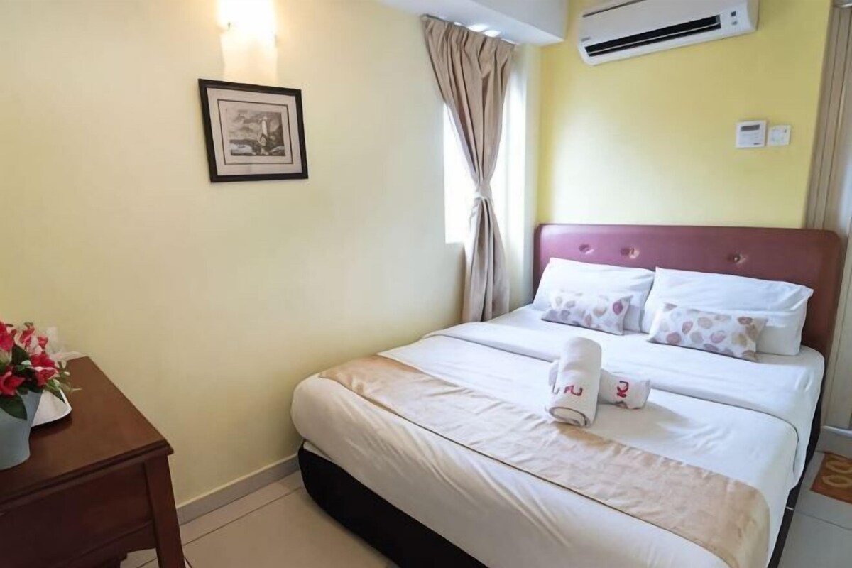 A hotel room with a bed, a bedside table with a vase of flowers, a wall-mounted air conditioner, and a framed picture on the wall.