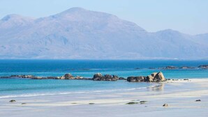 Een strand met helderblauw water, rotsformaties en bergen op de achtergrond.