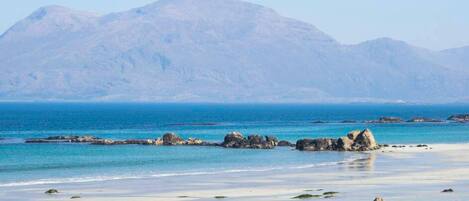 A beach with clear blue water, rocky outcrops, and mountains in the background.