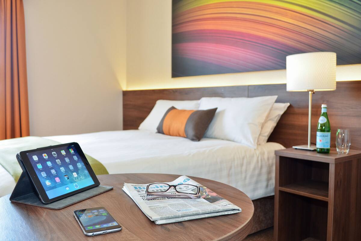 Habitación de hotel impecable con cama, mesita de noche, lámpara y una tableta sobre la mesita.