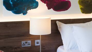 A hotel room with a bed, a nightstand with a lamp, a colorful patterned sofa, and a wall with abstract art.