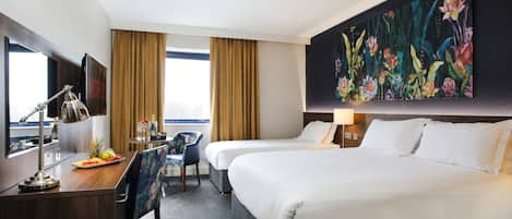 Habitación de hotel con dos camas, un escritorio con lámpara, una silla, una bandeja de frutas y un cuadro en la pared.