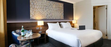 Ein Hotelzimmer mit einem großen Bett, zwei Nachttischlampen, einem kleinen runden Tisch mit Flasche und Gläsern, einem Sessel und einem Wandbild.