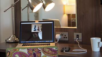 Een laptop op een bureau met een kleurrijke stoffen hoes, een witte mok, een lamp en een spiegel aan de muur.