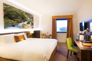 A hotel room with a large bed, a desk with a lamp, a chair, a small table with a bottle, and a painting of a waterfall on the wall. - Nox Hotel (Galway)
