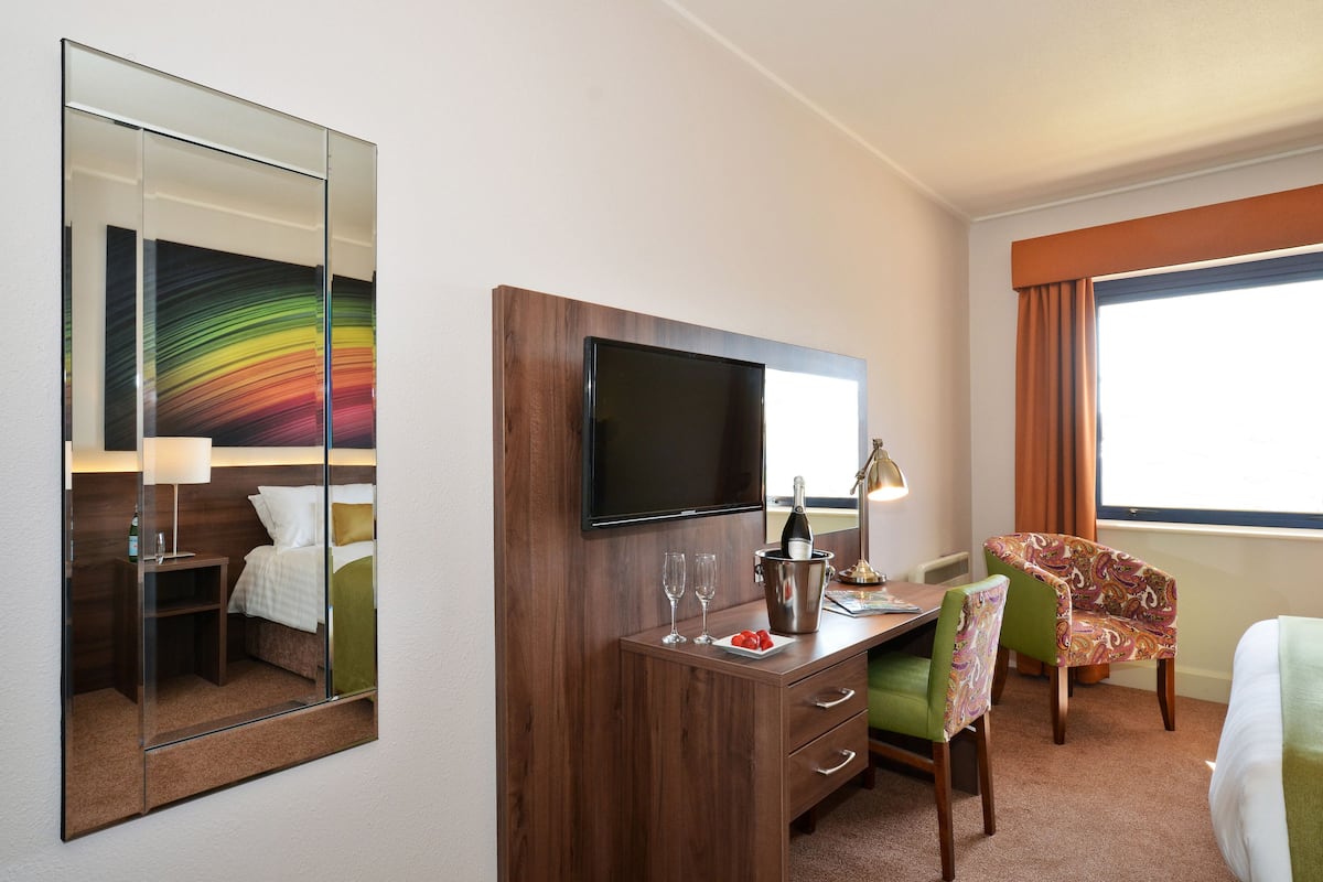Habitación de hotel con cama, escritorio con televisor de pantalla plana, silla, mesita con una botella de champán y espejo.