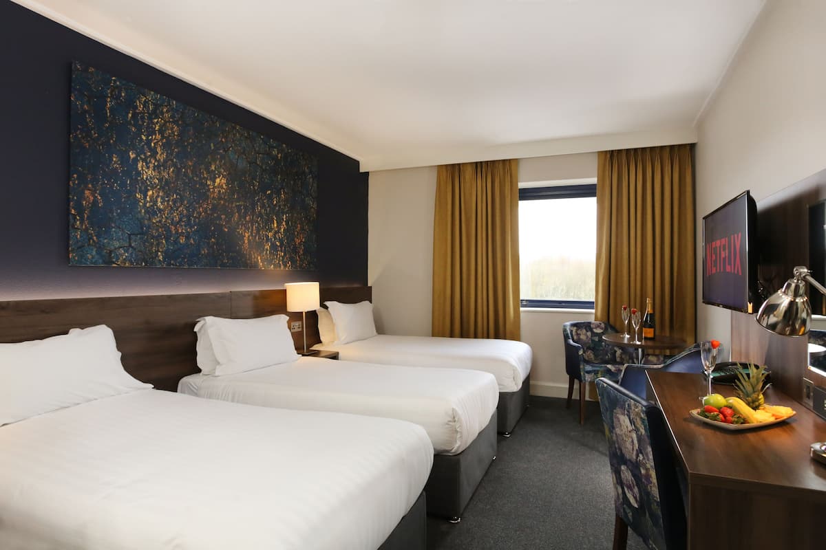 Habitación de hotel con tres camas, un escritorio con una bandeja de frutas, un televisor y un cuadro abstracto grande en la pared.
