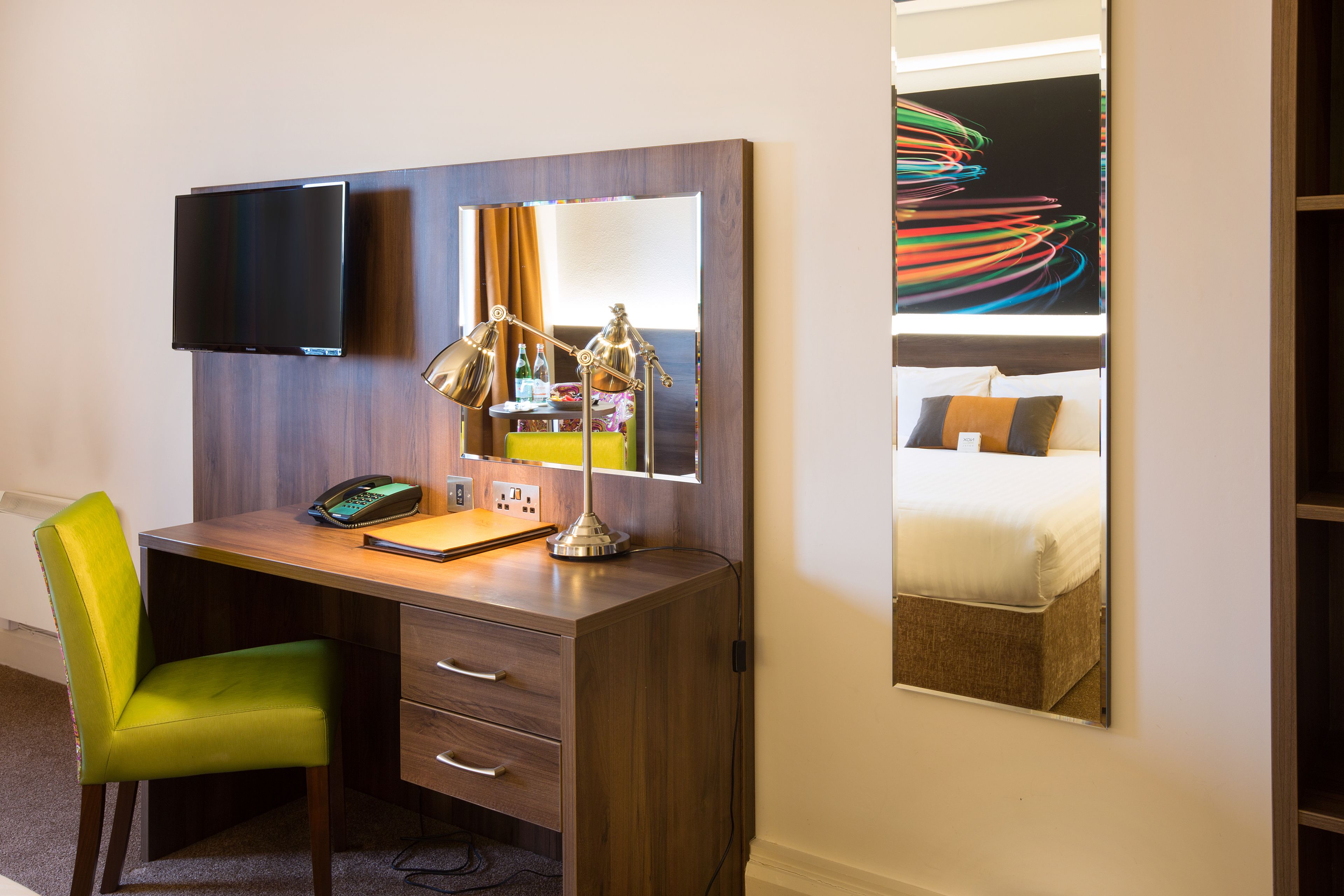 A hotel room with a wooden desk, a green chair, a flat-screen TV mounted on the wall, a desk lamp, a mirror, and a bed with a colorful abstract painting above it.