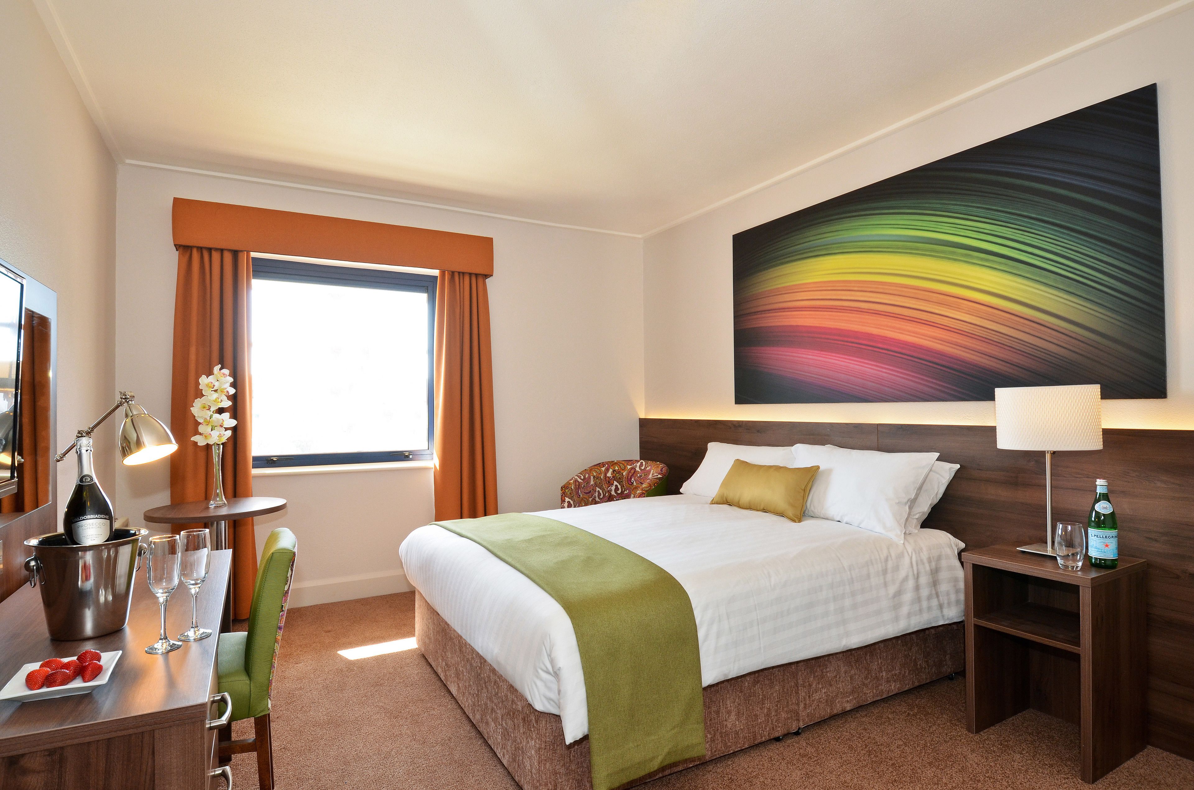 A hotel room with a bed, a desk with a bottle of champagne, a vase with flowers, and a colorful abstract painting on the wall.