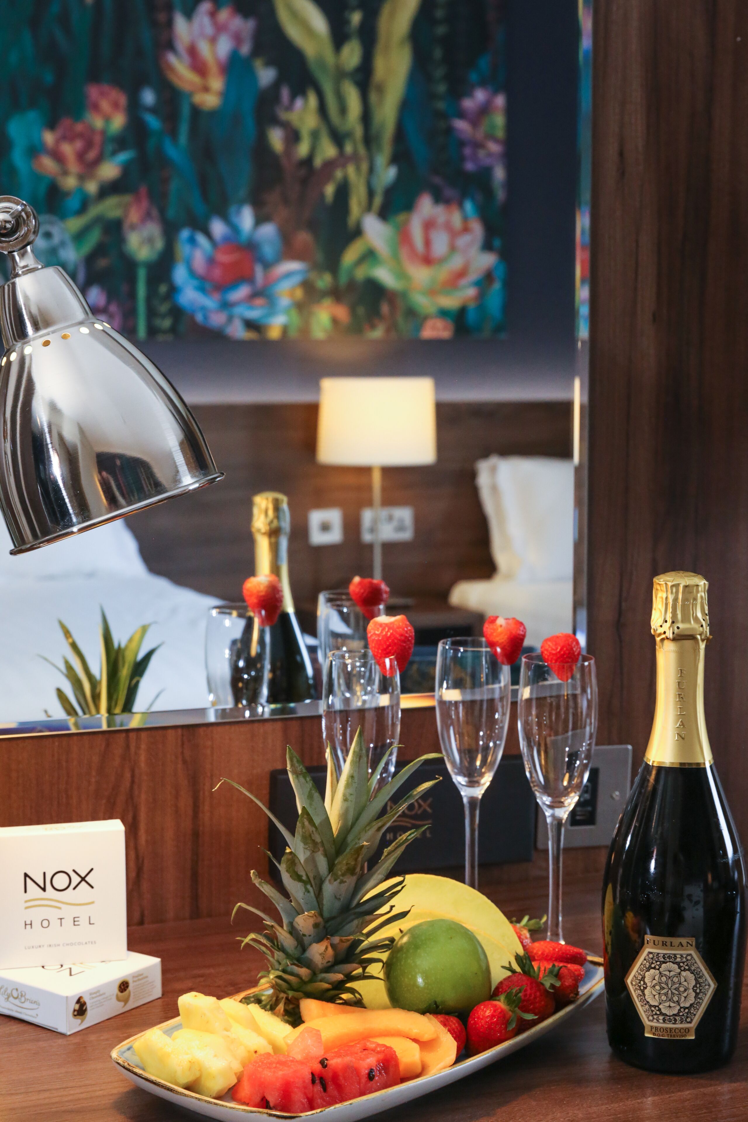 Une chambre d’hôtel avec un assortiment de fruits, une bouteille de champagne et un panneau indiquant « Nox Hotel ».