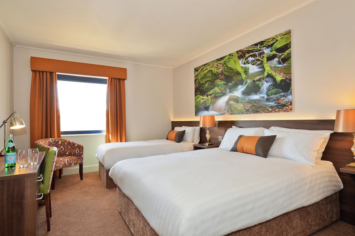Habitación de hotel con dos camas, un escritorio con una silla, una lámpara y un cuadro de una cascada en la pared.
