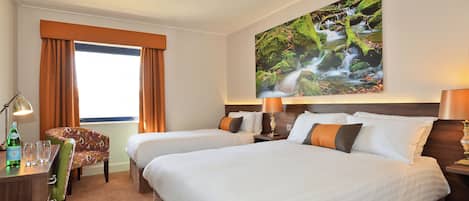 Habitación de hotel con dos camas, un escritorio con una silla, una lámpara y un cuadro de una cascada en la pared.