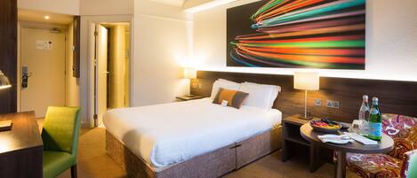 A hotel room with a bed, a desk, a chair, a table with bottles, and a colorful wall art piece.