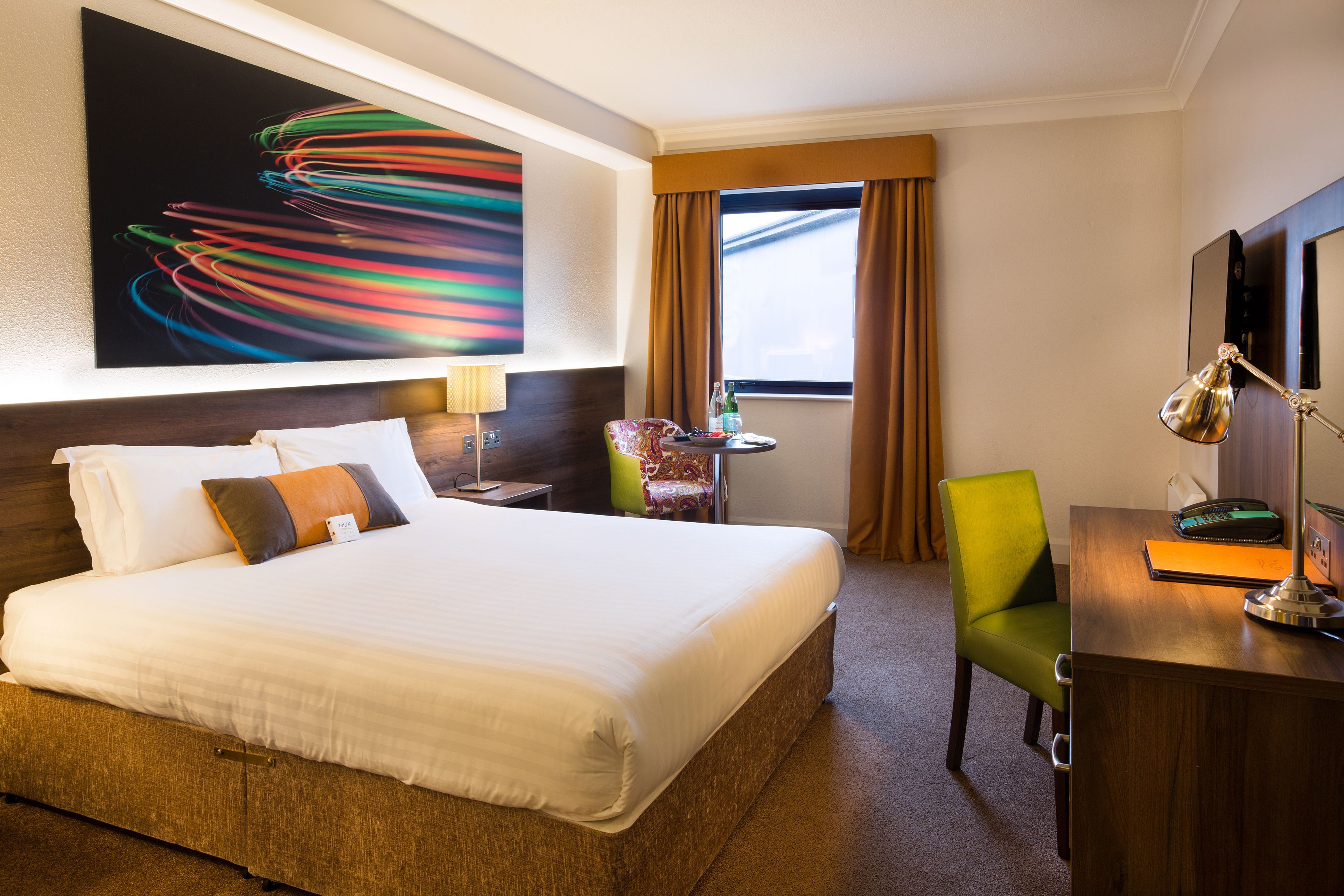 A hotel room with a large bed, a desk with a lamp, a chair, a window with curtains, and a colorful abstract painting on the wall.